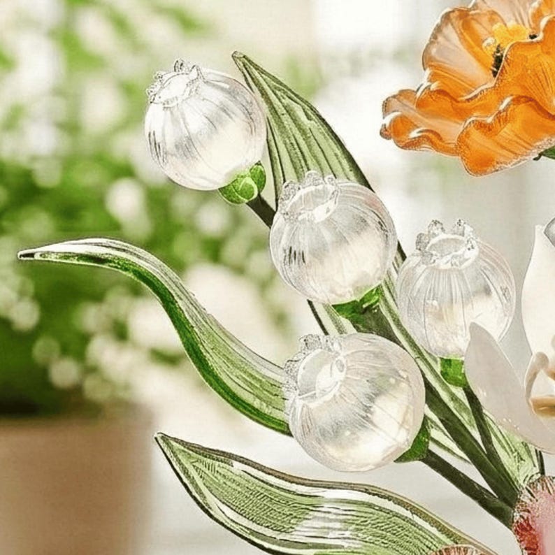 May include: Close-up of a glass floral arrangement featuring clear, spherical flower buds with textured details. The arrangement includes green leaves and a vibrant orange flower, set against a blurred background, creating a delicate and artistic display.
