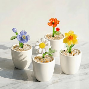 May include: A collection of five small, white ceramic pots, each containing a unique glass flower. The flowers are in various colors, including blue, orange, yellow, and white, with green stems and leaves. The pots are filled with small, light-colored pebbles.