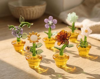 May include: A collection of colorful glass flower sculptures in small yellow pots. The flowers include a sunflower, a daisy, and other floral designs. The glass flowers are arranged on a wooden surface.