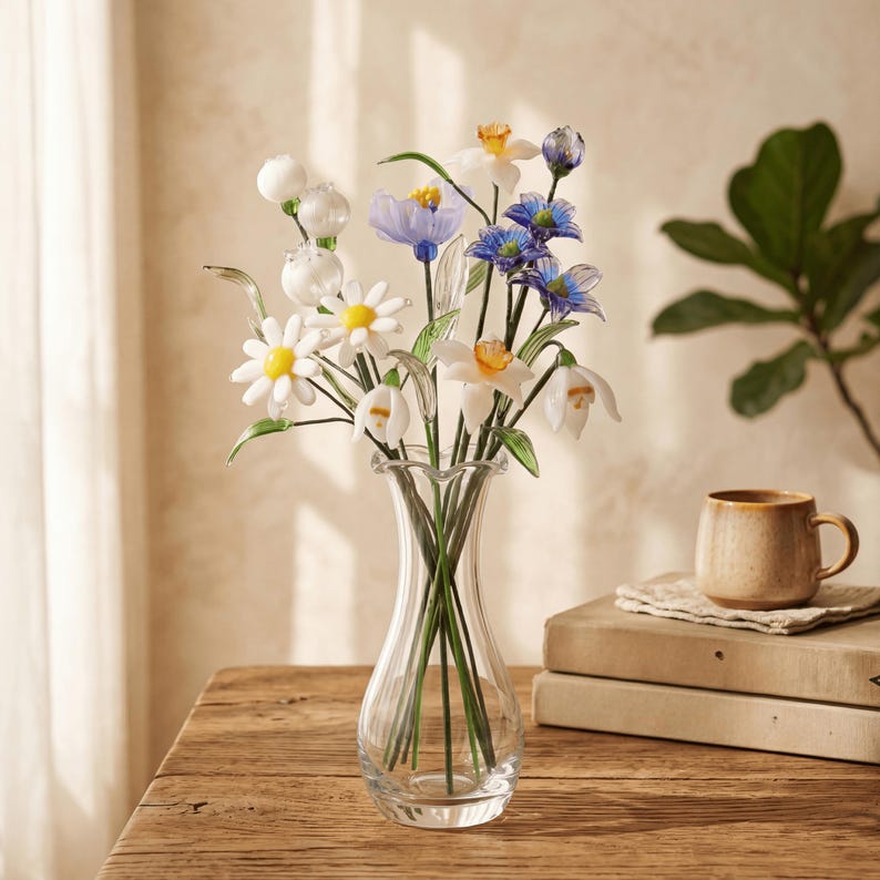 May include: A clear glass vase holds a bouquet of artificial flowers. The flowers include white daisies with yellow centers, blue and purple blooms, and white bell-shaped flowers. The vase sits on a wooden table, with a coffee cup and books in the background.