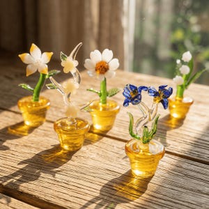 May include: A collection of five glass flower sculptures in small yellow pots. The flowers are various colors, including white, yellow, and blue, with green stems and leaves. The glass is transparent, and the pots are a solid yellow.
