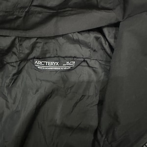 May include: Close-up of a black jacket interior with the Arc'teryx logo and size tag. The tag reads "XL/TG MEN'S/HOMMES" and "MADE IN VIETNAM/FABRIQUE AU VIETNAM". The jacket material appears to be a smooth, lightweight fabric.