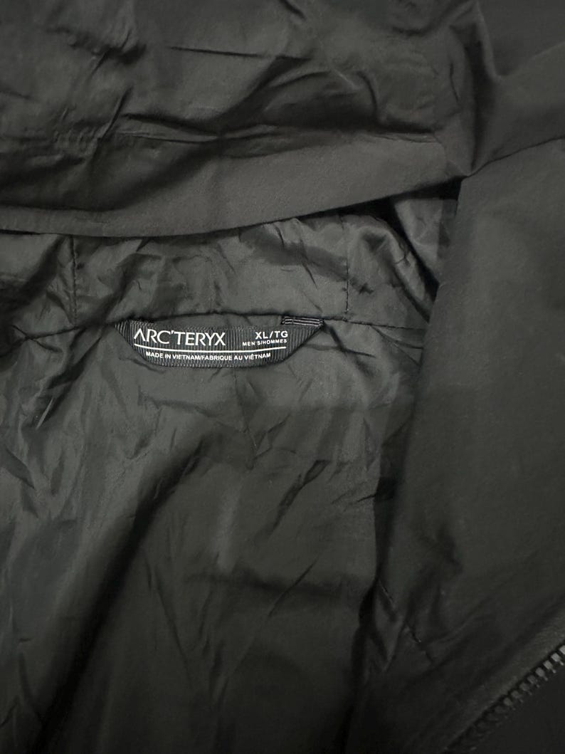 May include: Close-up of a black jacket interior with the Arc'teryx logo and size tag. The tag reads "XL/TG MEN'S/HOMMES" and "MADE IN VIETNAM/FABRIQUE AU VIETNAM". The jacket material appears to be a smooth, lightweight fabric.