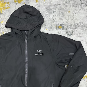 May include: Black hooded jacket with a full-length zipper and the Arc'teryx logo on the chest. The jacket has a hood and two zippered pockets. The GORE-TEX logo is visible on the sleeve. The jacket is made of a lightweight, waterproof material.