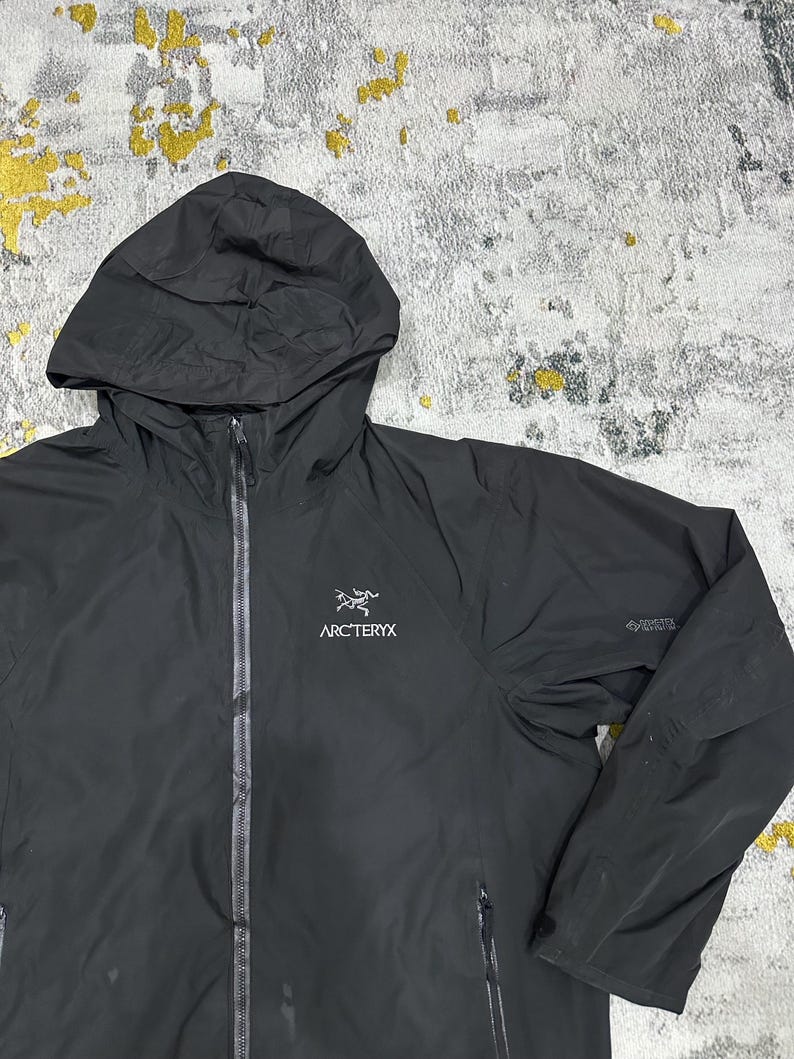 May include: Black hooded jacket with a full-length zipper and the Arc'teryx logo on the chest. The jacket has a hood and two zippered pockets. The GORE-TEX logo is visible on the sleeve. The jacket is made of a lightweight, waterproof material.