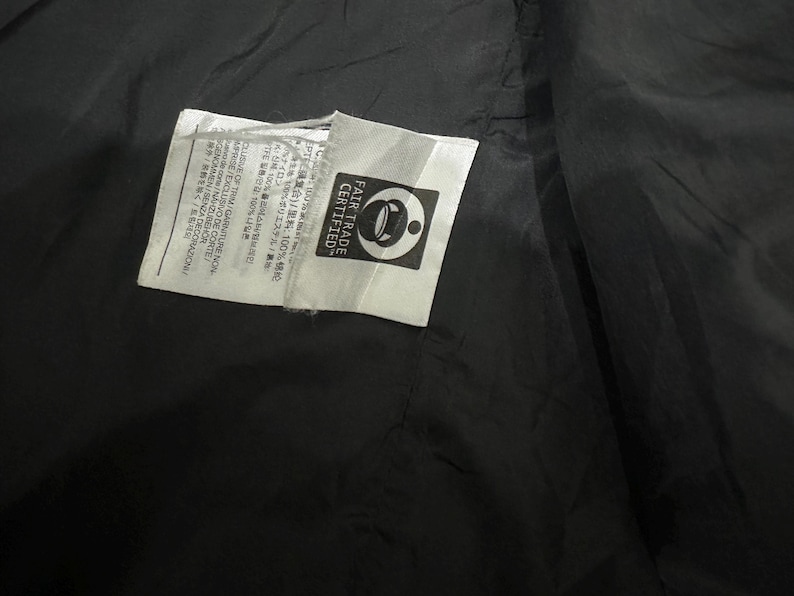 May include: Close-up of a black fabric with two white tags. One tag has text in multiple languages and the other tag has the words "FAIR TRADE CERTIFIED" and a logo.