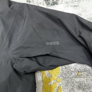 May include: Close-up of a dark gray jacket with a pocket and the GORE-TEX INFINIUM logo. The jacket appears to be made of a lightweight, water-resistant material. The image shows the texture and stitching details.