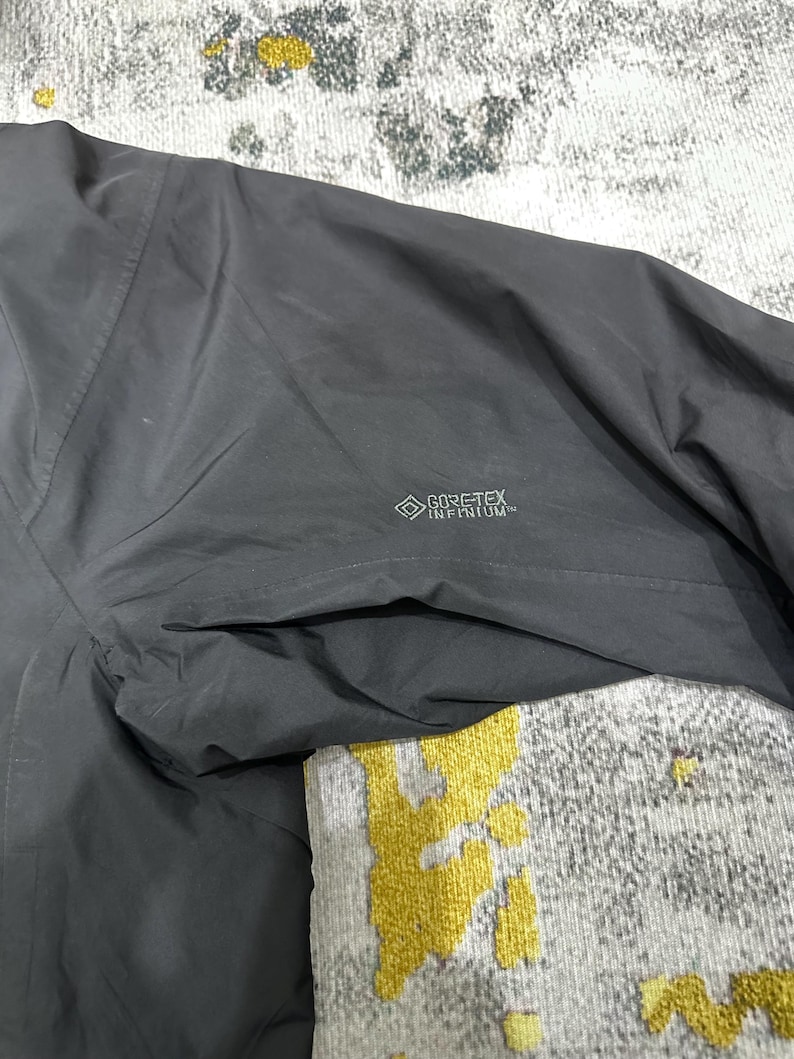 May include: Close-up of a dark gray jacket with a pocket and the GORE-TEX INFINIUM logo. The jacket appears to be made of a lightweight, water-resistant material. The image shows the texture and stitching details.