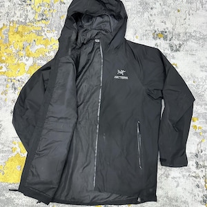 May include: Black hooded jacket with a full-length zipper and two side pockets. The jacket features the brand name "Arc'teryx" in white lettering on the chest. The jacket has a hood and is made of a lightweight material.