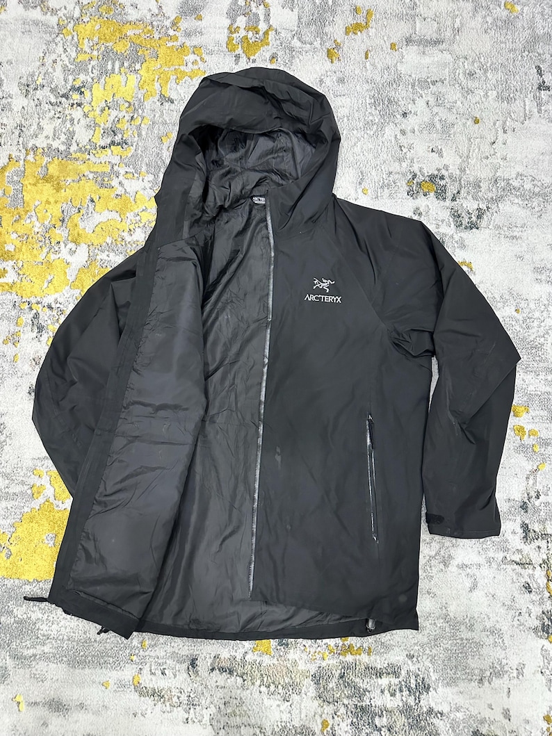 May include: Black hooded jacket with a full-length zipper and two side pockets. The jacket features the brand name "Arc'teryx" in white lettering on the chest. The jacket has a hood and is made of a lightweight material.
