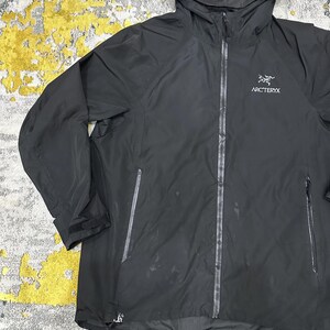 May include: Black hooded jacket with the brand name "Arc'teryx" printed on the chest. The jacket has a full-length zipper, two side pockets, and adjustable cuffs. The jacket is made of a lightweight, waterproof material.