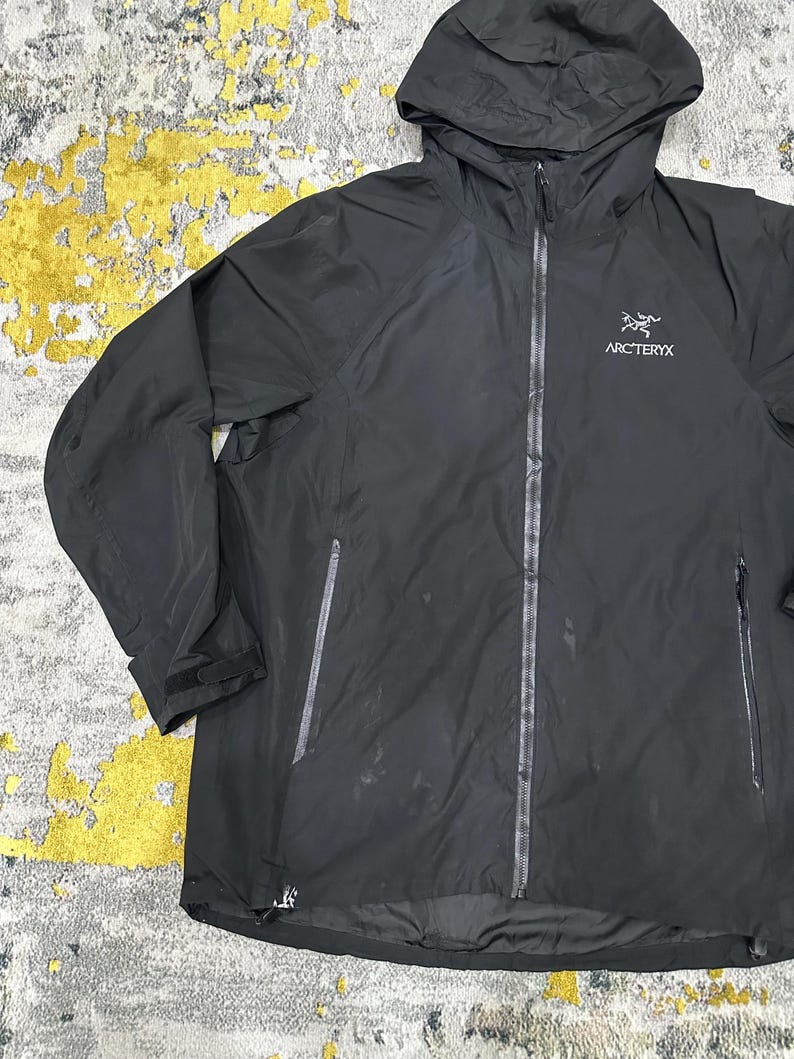 May include: Black hooded jacket with the brand name "Arc'teryx" printed on the chest. The jacket has a full-length zipper, two side pockets, and adjustable cuffs. The jacket is made of a lightweight, waterproof material.