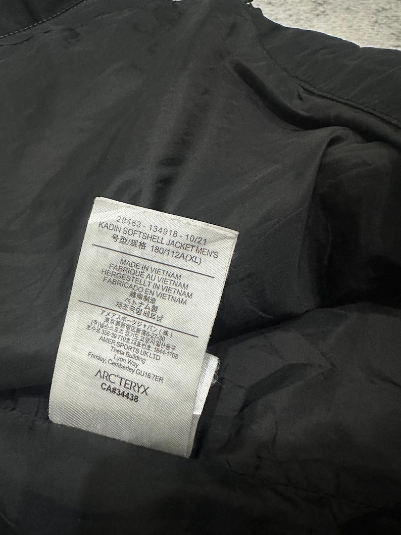 May include: A close-up of a black softshell jacket with a tag. The tag includes the text "KADIN SOFTSHELL JACKET MEN'S" and "MADE IN VIETNAM". The jacket is made in Vietnam.