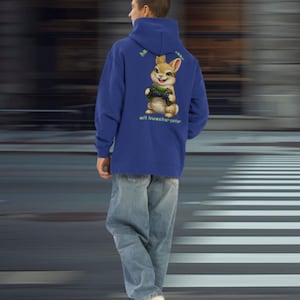 Könnte beinhalten: Ein königsblauer Hoodie mit einer Grafik eines Cartoon-Kaninchens, das einen Videospiel-Controller hält. Das Kaninchen ist braun und hellbraun mit grünen Akzenten. Der Hoodie hat den Text "he" und "mit knutsch-pfote" auf dem Rücken. Die Person trägt eine hellblaue Jeans und weiße Turnschuhe.