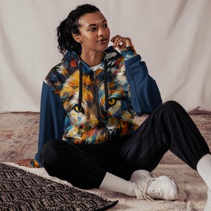 Könnte beinhalten: Ein Kapuzenpullover mit Reißverschluss und einem lebendigen, künstlerischen Löwengesicht-Design in Orange, Gelb, Blau und Grün. Der Hoodie hat marineblaue Ärmel und einen schwarzen Reißverschluss. Die Person trägt eine schwarze Hose und weiße Turnschuhe.