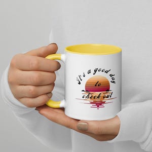 Könnte beinhalten: Weiße Keramik-Tasse mit gelbem Innenraum und Henkel. Die Tasse zeigt ein Sonnenuntergangsdesign mit dem Text "It's a good day to check out" in schwarzer Schrift. Die Tasse wird von einer Person gehalten.