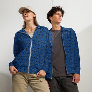 Puede incluir: Dos personas con chaquetas azules con cremallera a juego con un patrón geométrico. Las chaquetas tienen una cremallera blanca y un diseño repetitivo en tonos azules y marrones. Una persona lleva una gorra beige y pantalones color caqui, la otra una camiseta gris y vaqueros negros.