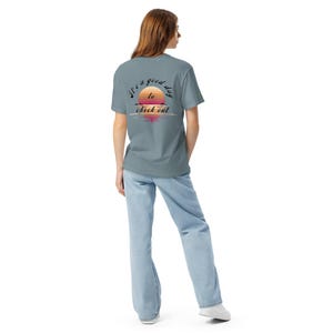 Könnte beinhalten: Hellblaues T-Shirt mit einem Sonnenuntergang-Motiv und dem Text "It's a good day to check out". Die Grafik zeigt eine Sonne mit orangefarbenen, rosa und gelben Farbtönen. Das Shirt besteht aus einem weichen Material und eignet sich für Freizeitkleidung.
