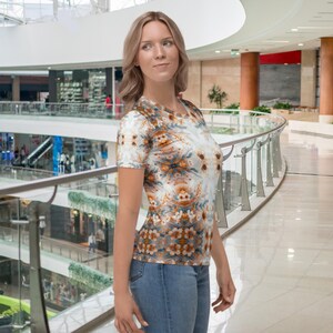 Könnte beinhalten: Ein kurzärmeliges T-Shirt mit Rundhalsausschnitt und einem Blumenmuster in Braun-, Orange- und Grautönen. Das Shirt wird mit einer blauen Jeans getragen. Der Hintergrund ist ein Einkaufszentrum mit einem geschwungenen Glasgeländer.