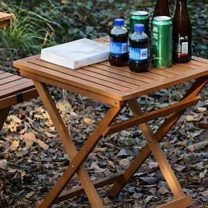 Può includere: Tavolino pieghevole in legno chiaro con piano a doghe. Sul tavolo ci sono bottiglie, lattine e un libro bianco. Il design delle gambe è a forma di X. Il tavolo è all'aperto.