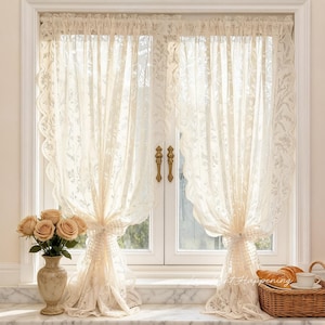 May include: Cream-colored lace curtains with scalloped edges frame a window. The curtains are tied back with matching fabric, revealing a glimpse of the outside. A vase of roses and a basket with pastries sit on the windowsill.