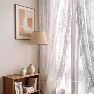 May include: A room interior featuring sheer white curtains with floral embroidery, a wooden bookshelf with books and vases, a floor lamp with a beige lampshade, and a framed abstract art piece on the wall. The curtains are tied back.