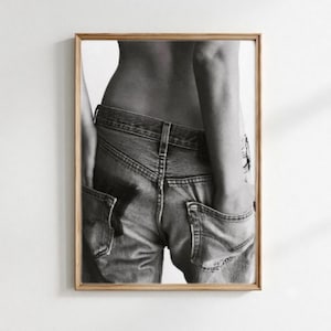 Può includere: Una fotografia in bianco e nero di un paio di jeans indossati bassi sui fianchi. I pantaloni in denim presentano due tasche posteriori e una cintura visibile, incorniciati da un semplice bordo in legno. L'immagine cattura la trama.