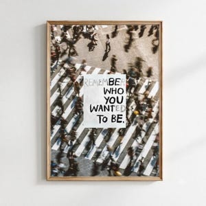 Könnte beinhalten: Ein rechteckiger Kunstdruck in einem hellen Holzrahmen mit einer bewegungsunscharfen Draufsicht auf Menschen, die einen Zebrastreifen überqueren. Ein weißes rechteckiges Feld in der Mitte zeigt den schwarzen Text: REMEMBER WHO YOU WANTED TO BE.