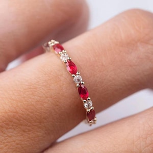May include: A gold band ring featuring alternating oval-cut red gemstones and round, clear gemstones. The ring is set with multiple gemstones, creating a continuous pattern around the band. The ring is shown on a person's finger.