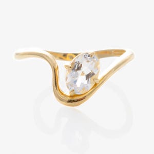 May include: A gold ring with a clear, oval-cut gemstone. The ring's band has a unique, flowing design. The gemstone is set in a prong setting, and the ring is displayed against a white background. The ring is a piece of jewelry.