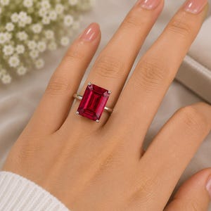 May include: A silver ring featuring a large, rectangular, emerald-cut deep red gemstone set in a four-prong setting. The band is thin and polished, showcasing the vibrant, saturated color of the stone against the metallic silver frame.