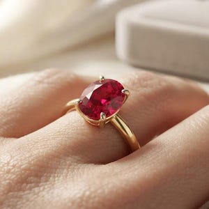 Peut inclure: Une bague en or ornée d'une grande pierre précieuse rouge vif de taille ovale, maintenue par un sertissage à quatre griffes. L'anneau est lisse et poli, présentant un design classique qui met en valeur la couleur cramoisie et la surface facettée.