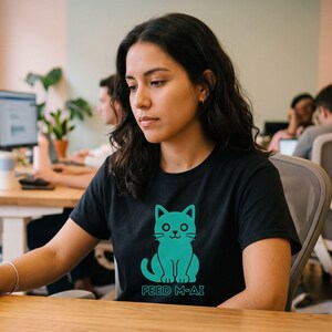 Puede incluir: Una camiseta negra de manga corta con un gráfico de color verde azulado de un gato sentado con ojos grandes y bigotes. Debajo de la ilustración del gato, el texto FEED M-AI está impreso en un color verde azulado a juego. Cuello redondo.
