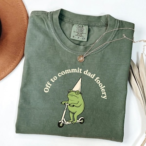 May include: Olive green t-shirt with the phrase "Off to commit dad foolery" in a semi-circle above a cartoon frog riding a scooter. The frog wears a party hat and glasses. A gold necklace is draped across the shirt.