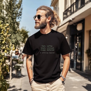 May include: Black crew neck t-shirt with green text: "fuel: coffee, level: critical, sleep: deferred, // refilling required." The shirt is worn by a person with sunglasses and a beard, standing outdoors.
