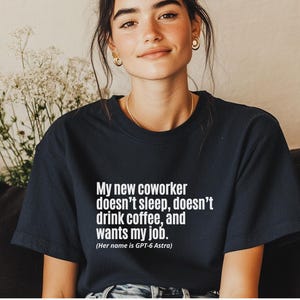 May include: A dark navy blue short-sleeved t-shirt featuring white text that reads: My new coworker doesn't sleep, doesn't drink coffee, and wants my job. (Her name is GPT-6 Astra). The shirt has a crew neckline and a standard fit.