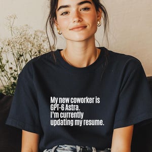 May include: A dark navy blue short-sleeved t-shirt featuring white text that reads: My new coworker is GPT-6 Astra. I'm currently updating my resume. The shirt has a classic crew neckline and a standard fit, providing a clean and simple appearance.