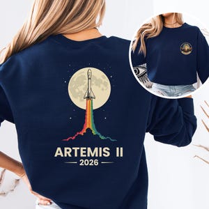 May include: Navy blue sweatshirt featuring a graphic of a rocket launching from a moon, with a rainbow trail. The text "ARTEMIS II - 2026" is printed below the graphic. A smaller graphic is on the upper right.