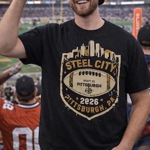 May include: Black t-shirt with a distressed gold and white graphic featuring the words "Steel City", a football, and the text "Draft 26 Pittsburgh 2026 PA". The shirt is worn by a person at a football game.