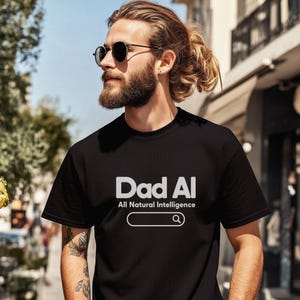 May include: A black short-sleeved t-shirt featuring the white text Dad AI, the phrase All Natural Intelligence, and a graphic of a search bar with a magnifying glass icon. The design is centred on the chest of the garment in a clean font.