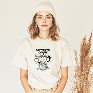 Könnte beinhalten: Cremefarbenes T-Shirt mit dem Text "SAY YES TO COFFEE" und einer schwarz-weißen Grafik einer lächelnden Kaffeemaschine, die eine Tasse Kaffee hält. Die Person im Bild trägt eine cremefarbene Mütze.