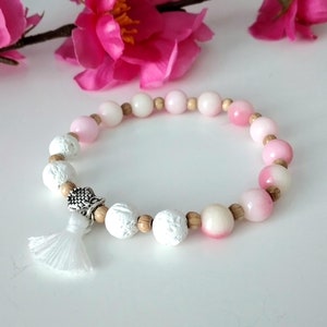 Può includere: Un braccialetto di perline rosa e bianche con un fiocco bianco e un ciondolo argentato. Il braccialetto è realizzato con legno, roccia lavica e perline rosa e bianche.