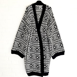 May include: A long, open-front cardigan in a black and white geometric pattern. The sweater has a shawl collar and wide sleeves with black trim. The knitwear is displayed against a white brick wall.