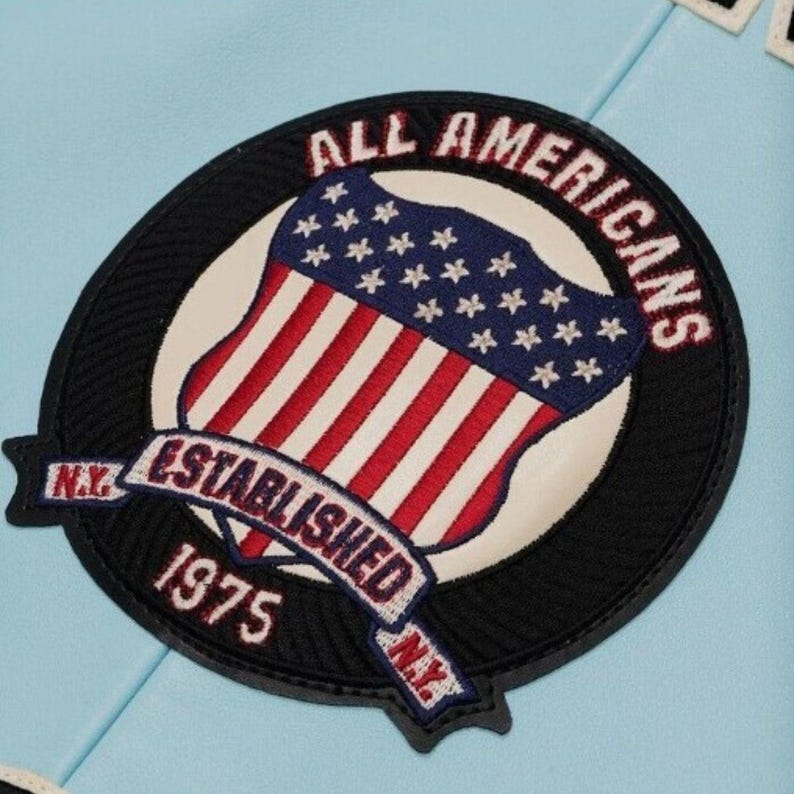 Może przedstawiać: Okrągła naszywka z napisem "ALL AMERICANS" w kolorze czerwonym, wygiętym u g&oacute;ry. Naszywka przedstawia wz&oacute;r tarczy z flagą amerykańską i napisem "ESTABLISHED 1975 N.Y." na banerze. Naszywka jest czarna z białymi i czerwonymi akcentami.