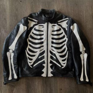 Könnte beinhalten: Schwarze Lederjacke mit weißem Skelettmuster. Die Jacke hat einen durchgehenden Reißverschluss und einen klassischen Kragen. Das Skelettmuster ist auf die Vorderseite und die Ärmel gemalt und sorgt für einen einzigartigen Look. Die Jacke liegt flach auf einer Holzoberfläche.