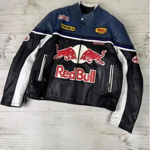 Puede incluir: Una chaqueta de moto de cuero en negro, azul y blanco con un destacado logotipo de Red Bull en rojo y blanco en el pecho. La chaqueta incluye varios parches de patrocinadores como Motul, Michelin, Bell Helmets y Ducati, con paneles laterales blancos.