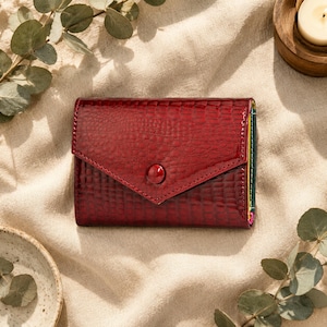 May include: A deep red wallet with a glossy, textured crocodile-skin pattern and a triangular envelope-style flap secured by a single matching red button. The side edges reveal thin, vertical stripes of green, yellow, and pink leather accents.