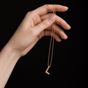 Puede incluir: Un collar dorado con un colgante con la letra "L". El colgante tiene un diseño de letra de bloque simple. El collar se sostiene sobre un fondo negro.