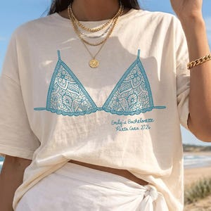 May include: A cream t-shirt featuring a turquoise lace bra graphic and the text "Emily's Bachelorette Punta Cana 2024". The outfit includes white wrap trousers, gold jewellery, and a straw hat, with a beach scene in the background.