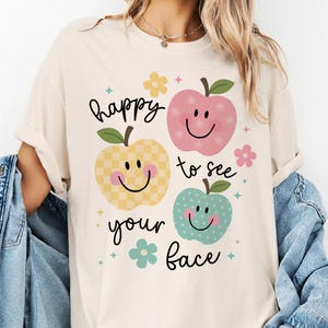 Op de afbeelding: Crèmekleurig T-shirt met de tekst "happy to see your face" en lachende appelafbeeldingen in roze, geel en blauw. Het shirt heeft een relaxte pasvorm en opgerolde mouwen. Een spijkerjasje is over de arm gedrapeerd.
